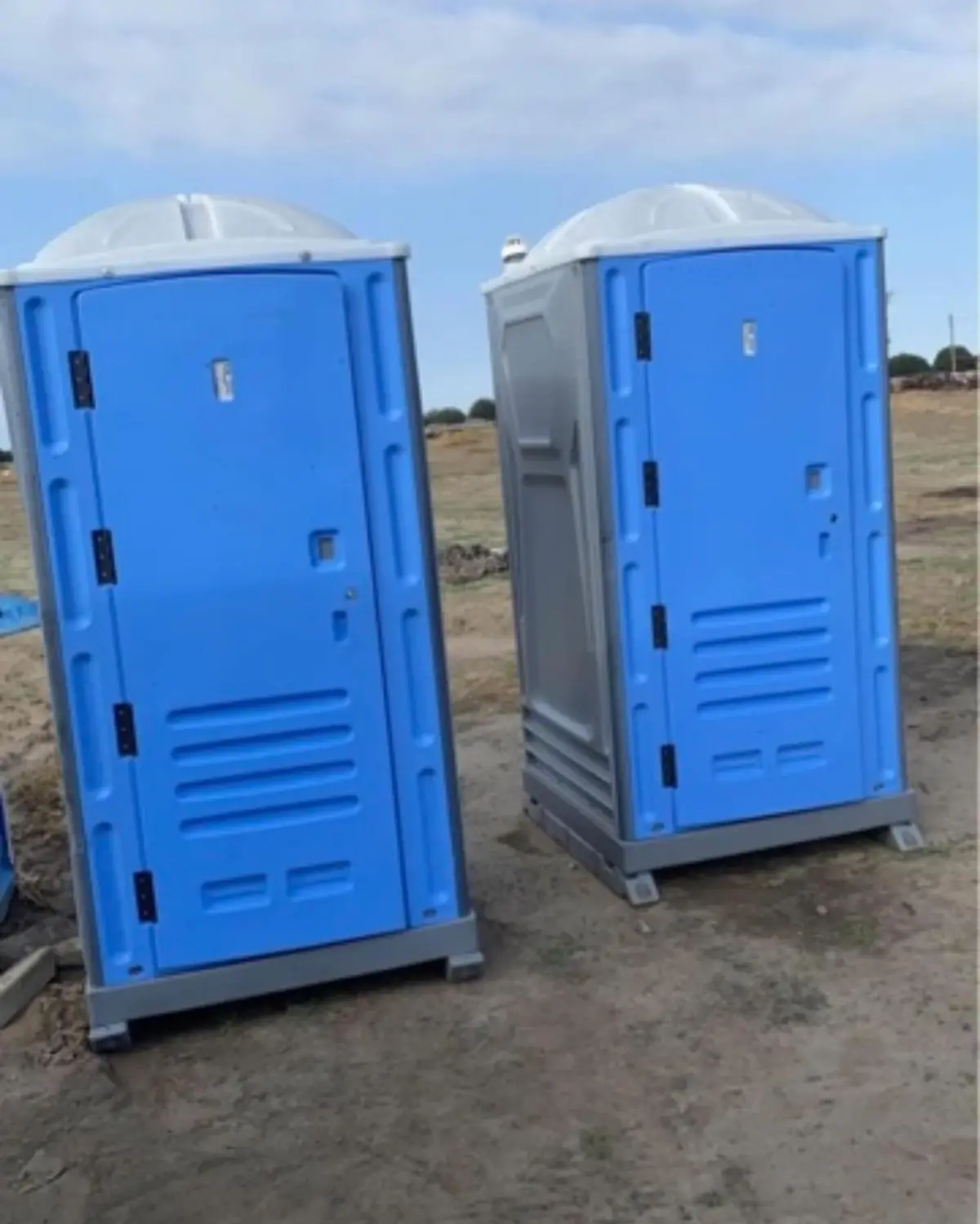 Two blue standard porta potties set on a dirt jobsite under a clear sky